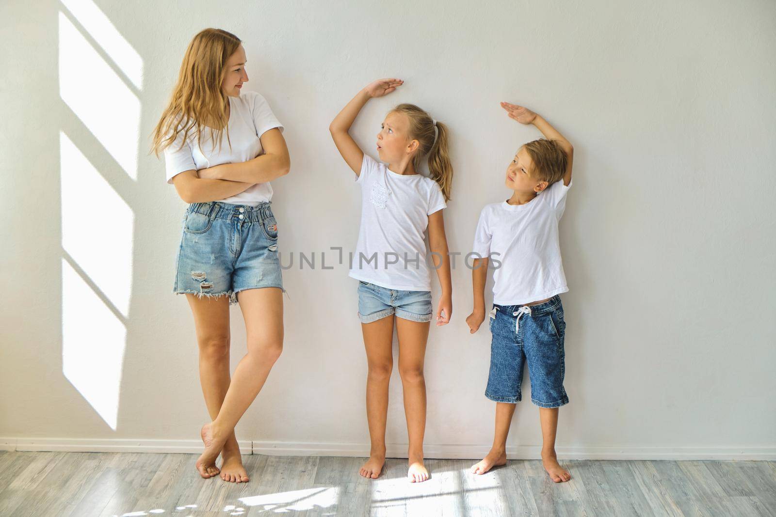 mother measures growth of kids to a daughter and son near light grey wall by InnaVlasova