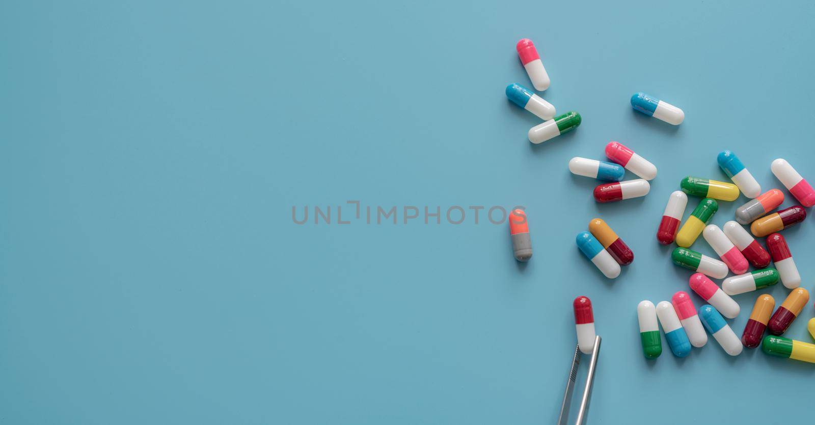 Antibiotic drug selection. Multi-colored capsule pills on blue background. Forceps picks a red-white capsule up from many antibiotic capsule pills. Chance of being selected. Probability sampling. by Fahroni