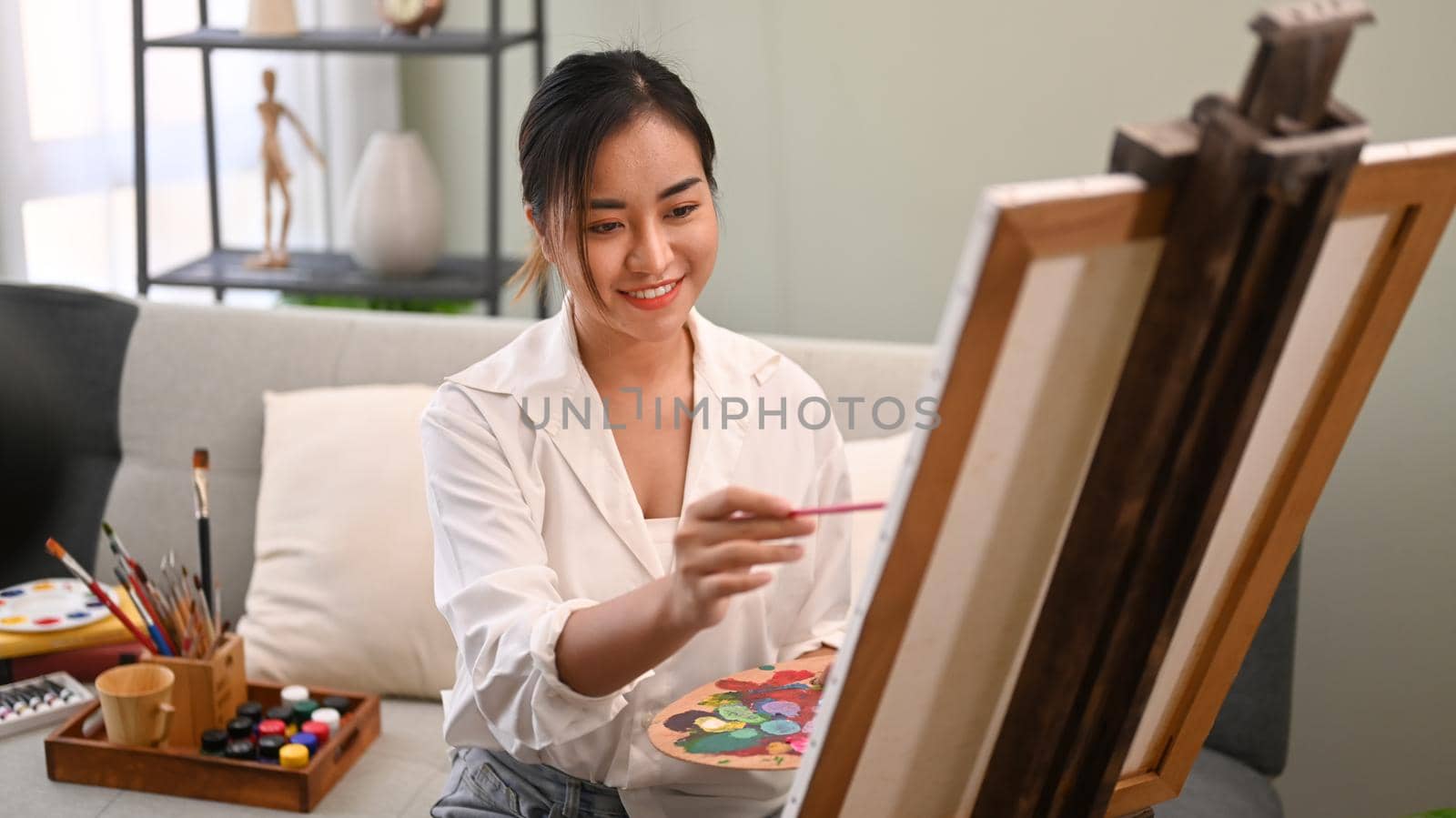 Smiling young woman painting picture on canvas with oil paints in bright cozy living room. Art, creative hobby and leisure activity concept by prathanchorruangsak