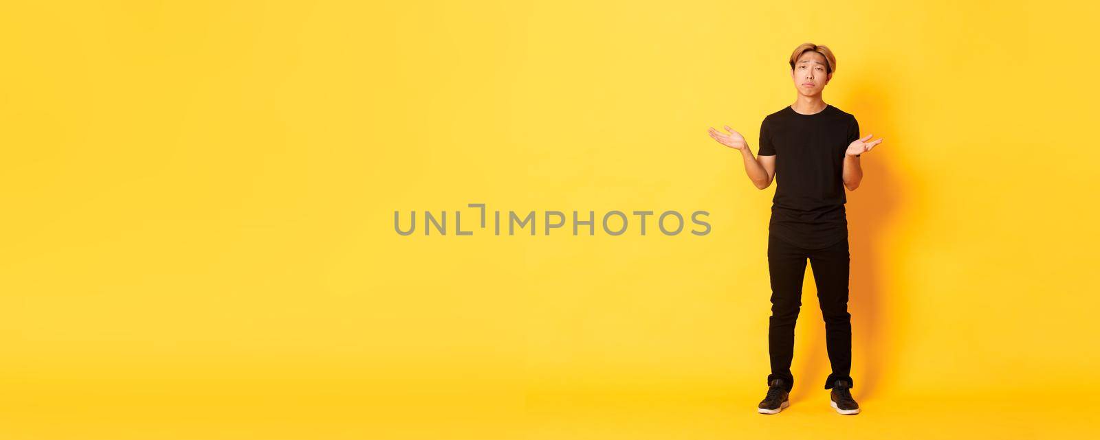 Full-length of disappointed and frustrated asian guy shrugging, looking sad, yellow background.