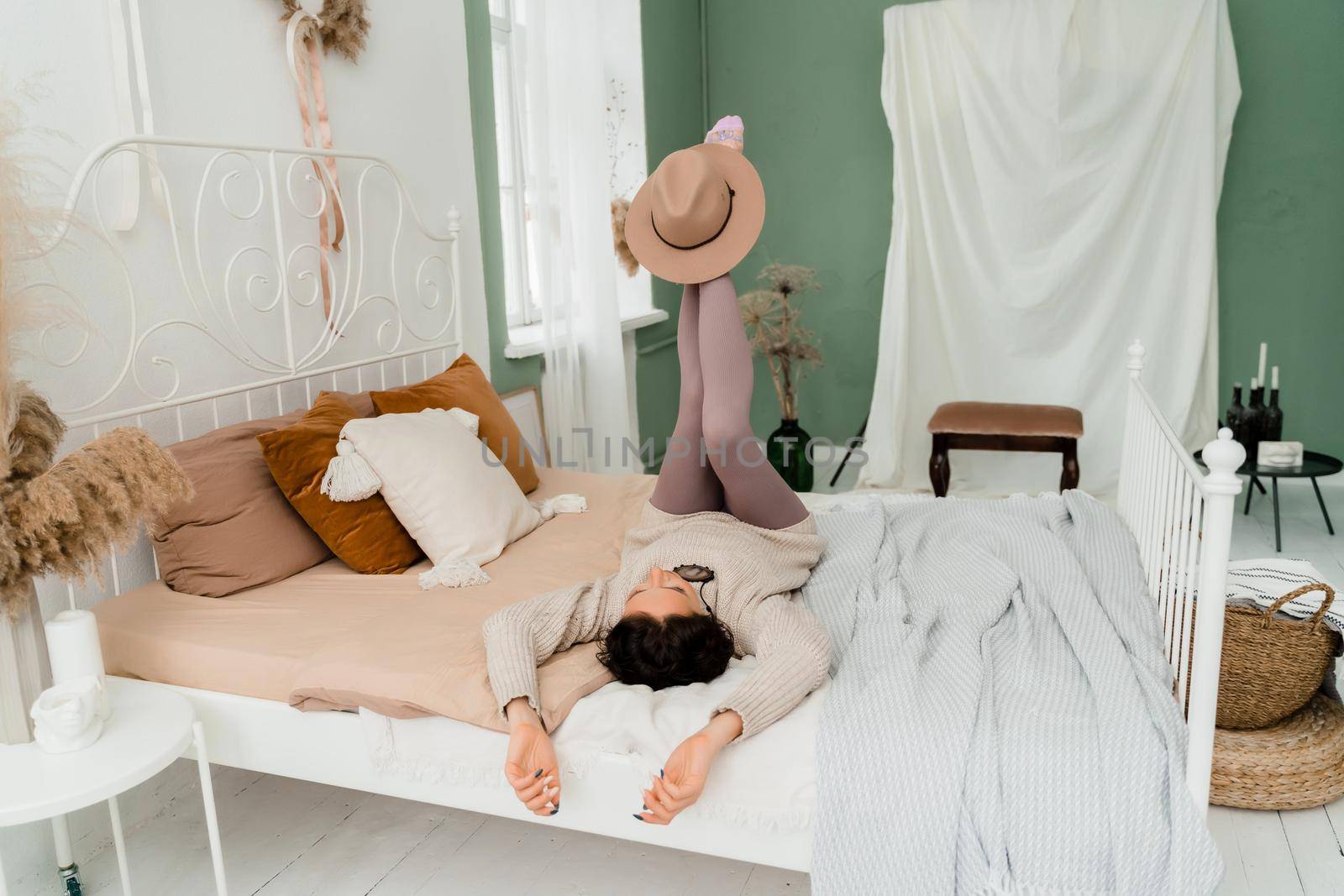 Lifestyle concept. A young middle-aged woman in a sweater lies on the bed on her back, laughs and holds a brown hat on her feet