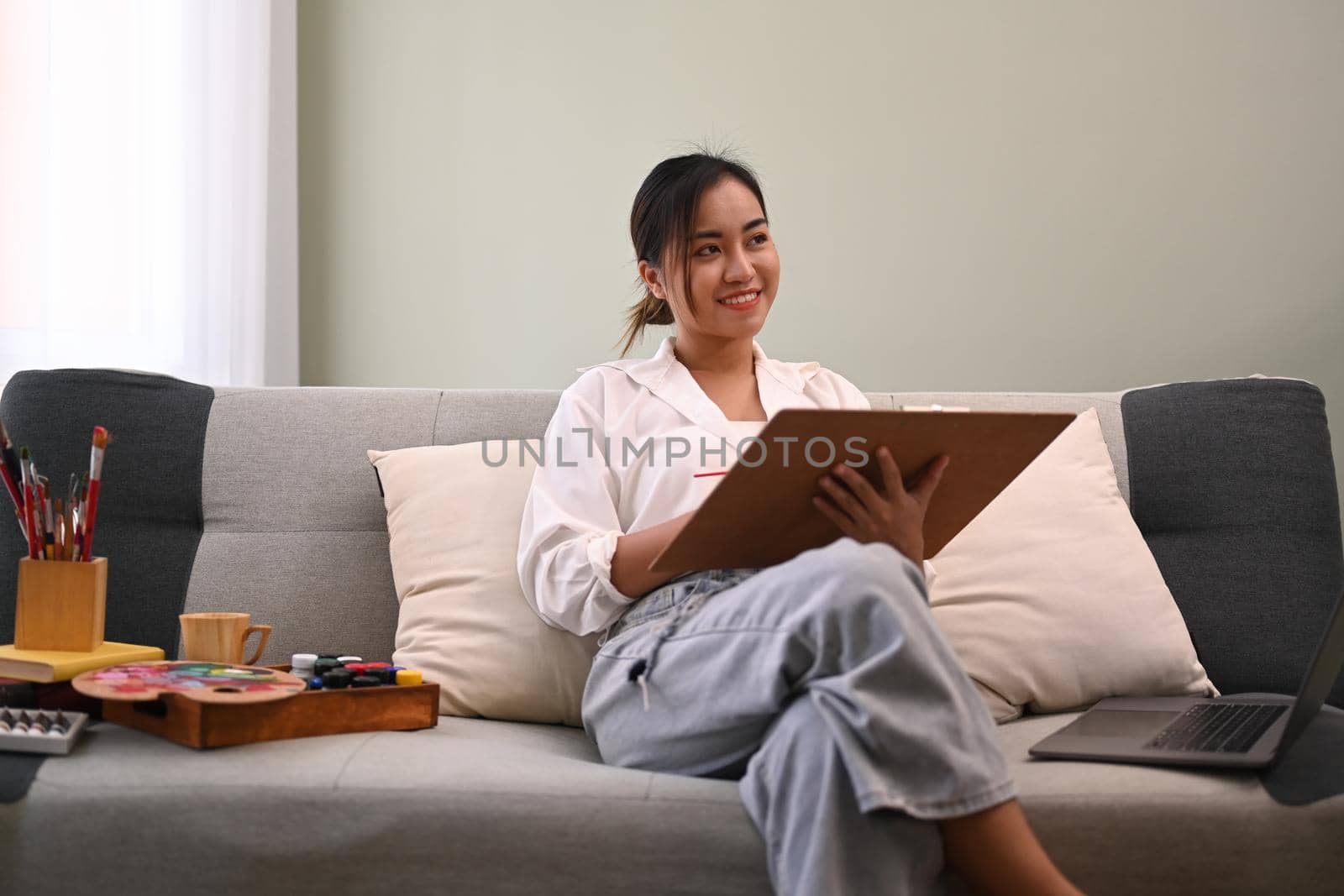 Peaceful asian woman paints picture with watercolor on couch at cozy home. Art, creative hobby and leisure activity concept.