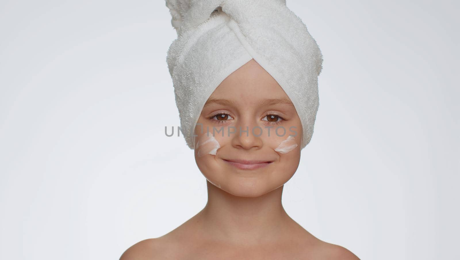 Lovely young child girl after bath in towel on head applying cleansing moisturizing cream. Teenager kid face skincare healthy treatment, natural creme cosmetics. Female portrait. Perfect fresh clean