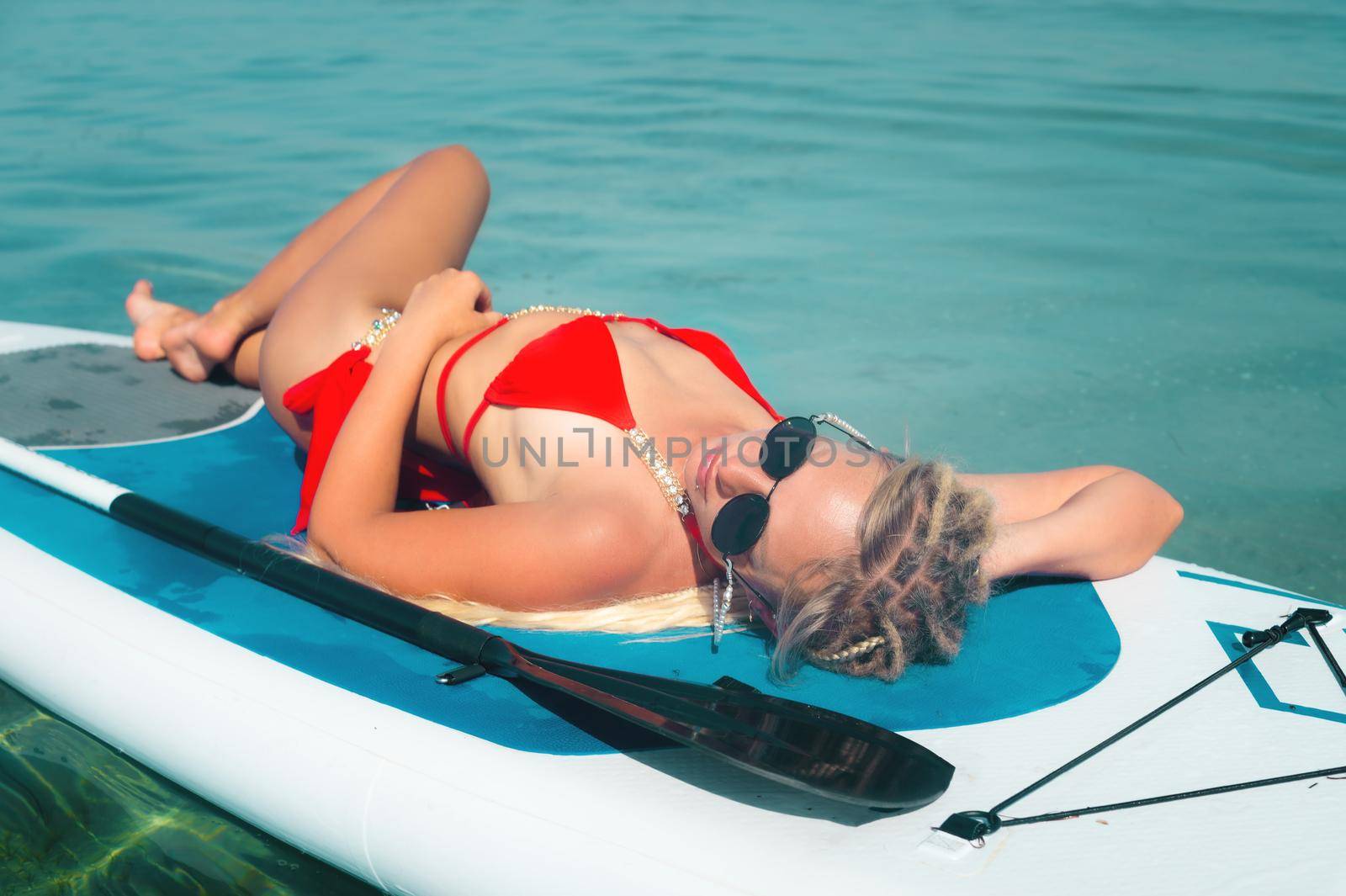 Young sexy surfer woman in sunglasses and bikini swimsuit lying on her board looking up at the sky. Rest on the sea.