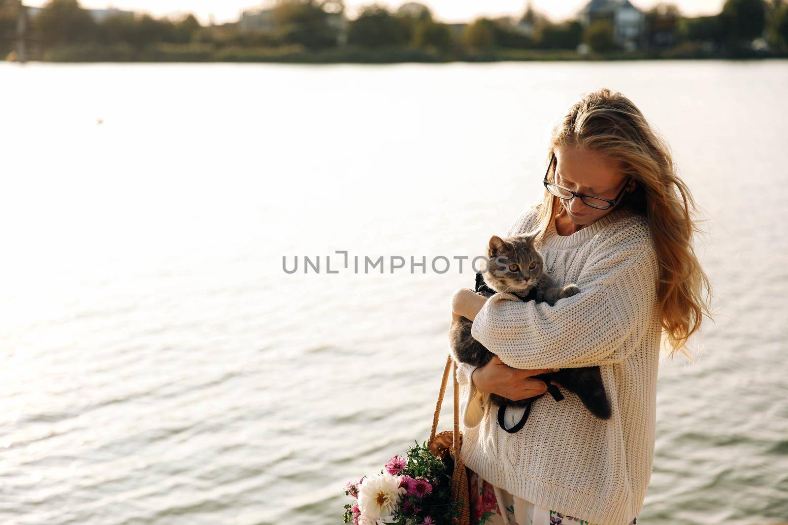 Young blonde, girl, woman with glasses holds a cat breed Scottish Straight. Walk with pet near the lake, river at sunset on the beach in Spring