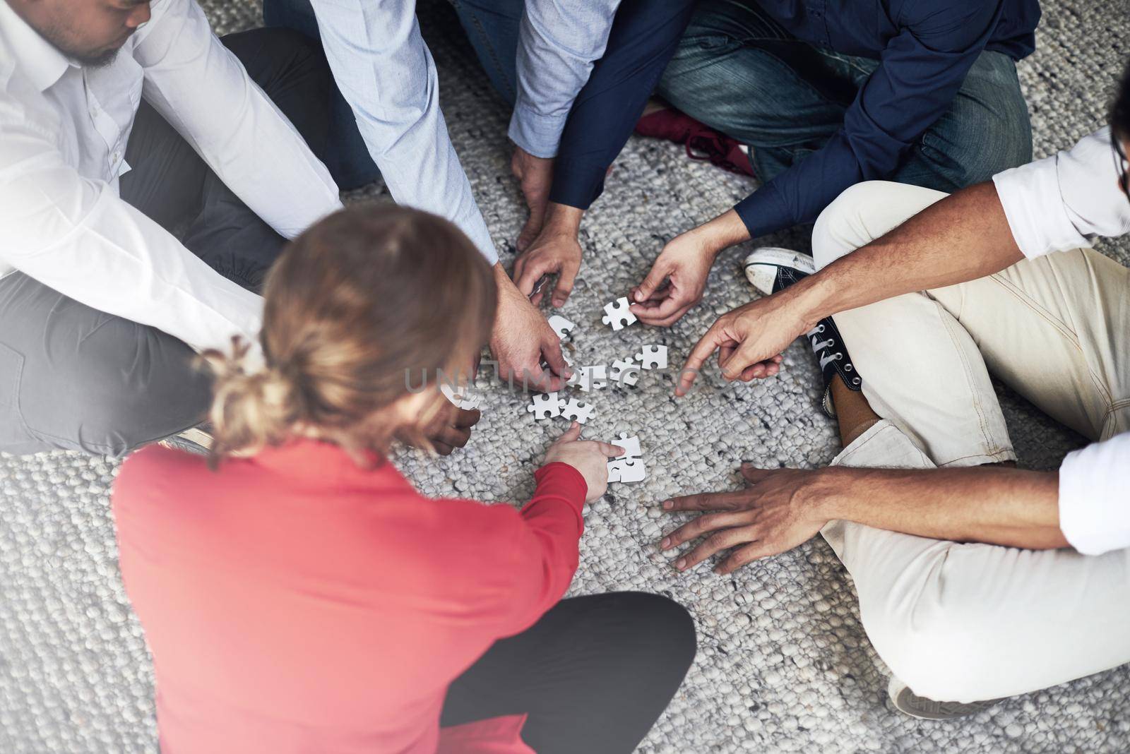 They love it when a plan comes together. High angle shot of a group of unidentifiable businesspeople building a puzzle together while sitting on a carpet in the office. by YuriArcurs