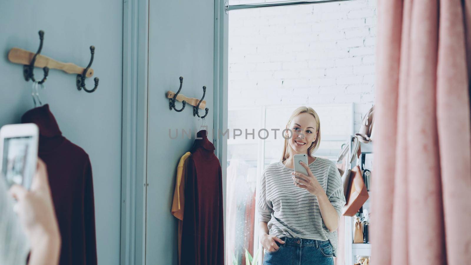 Pretty blond woman is making mirror selfie with smart phone while standing in nice fitting room in luxurious clothes boutique. She is posing, moving and smiling carelessly.