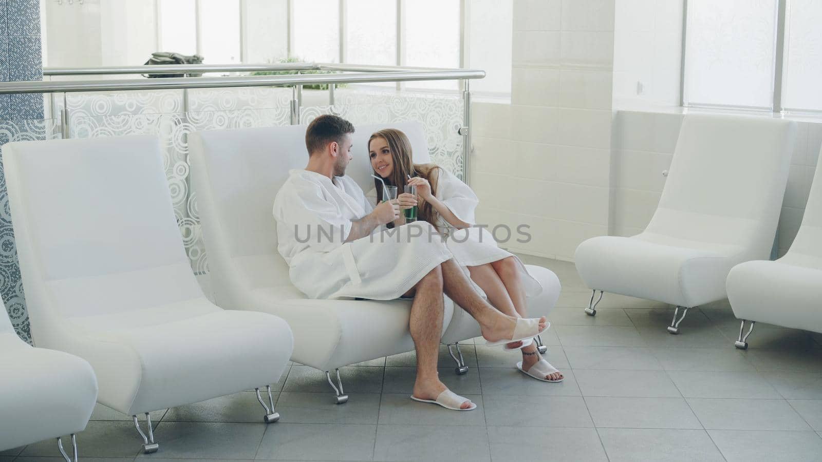 Young happy married couple is relaxing sitting in modern spa salon with cocktail glasses and chatting. Romantic relationship, relaxation and wellness concept.