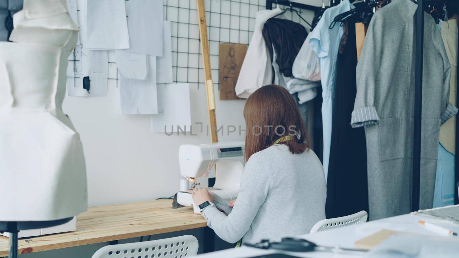 Back view of young woman clothing designer working with sewing machine sitting at studio desk. Sewing items, tailoring dummy and women's clothes are visible. by silverkblack