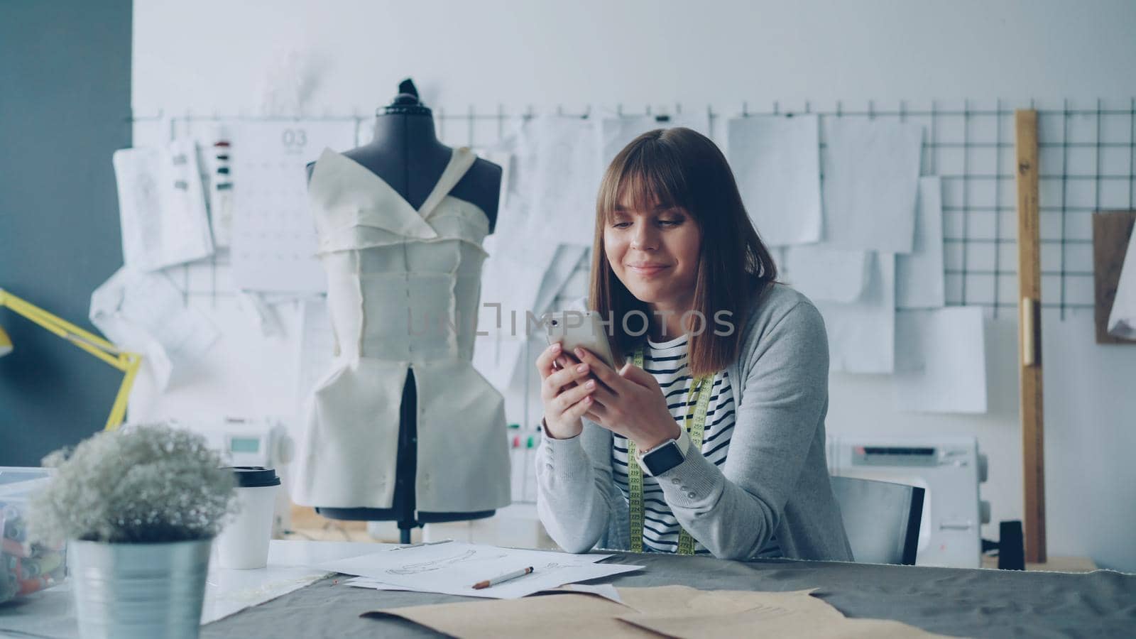 Creative clothing designer is looking at smartphone screen and drawing sketch while working in modern tailor shop at sewing table. Woman is busy and involved in process.