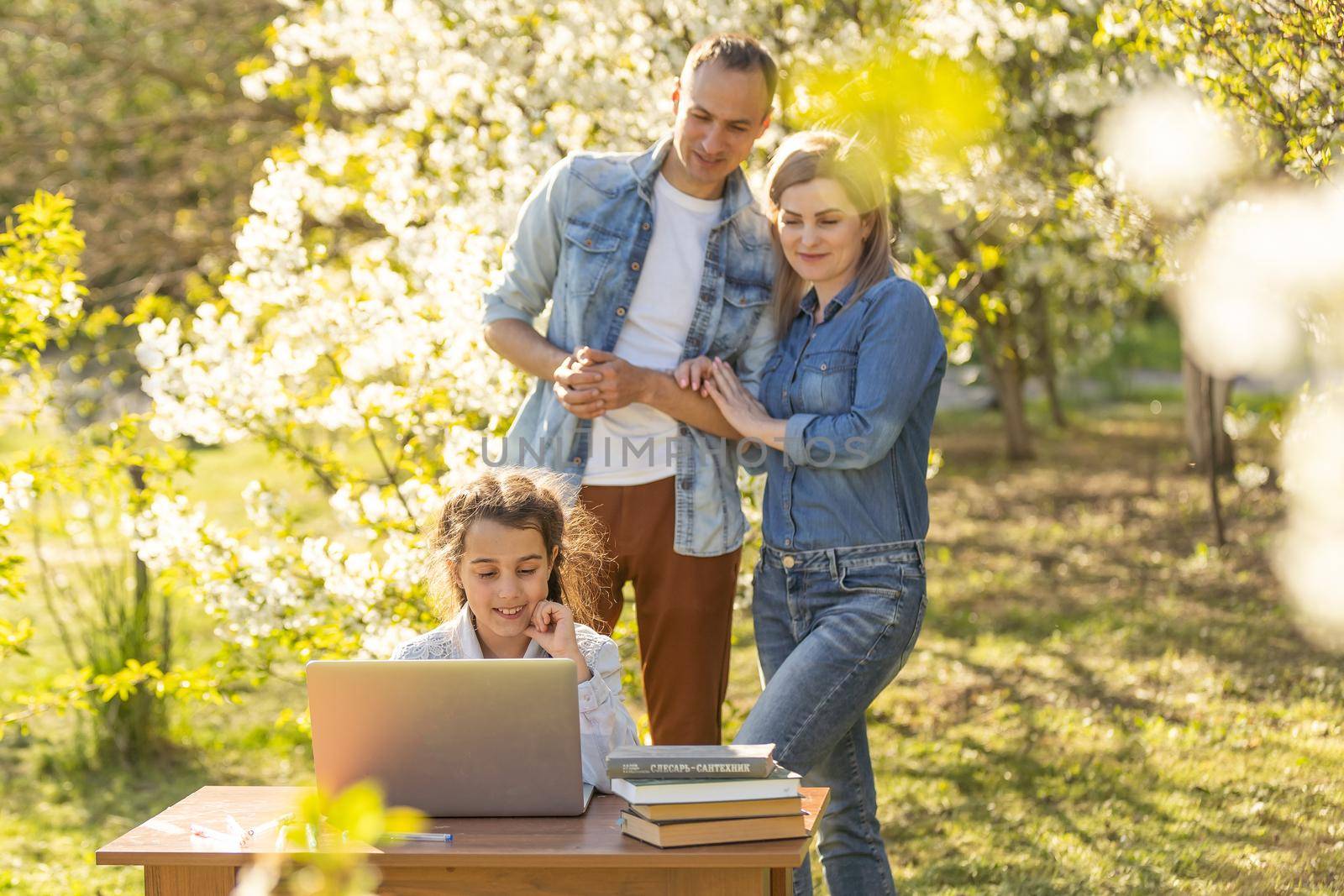 Happy family of four parents and cute little girl studying on laptop, enjoy using laptop watching cartoons, make internet video call or shopping online looking at computer screen sit together outdoor.