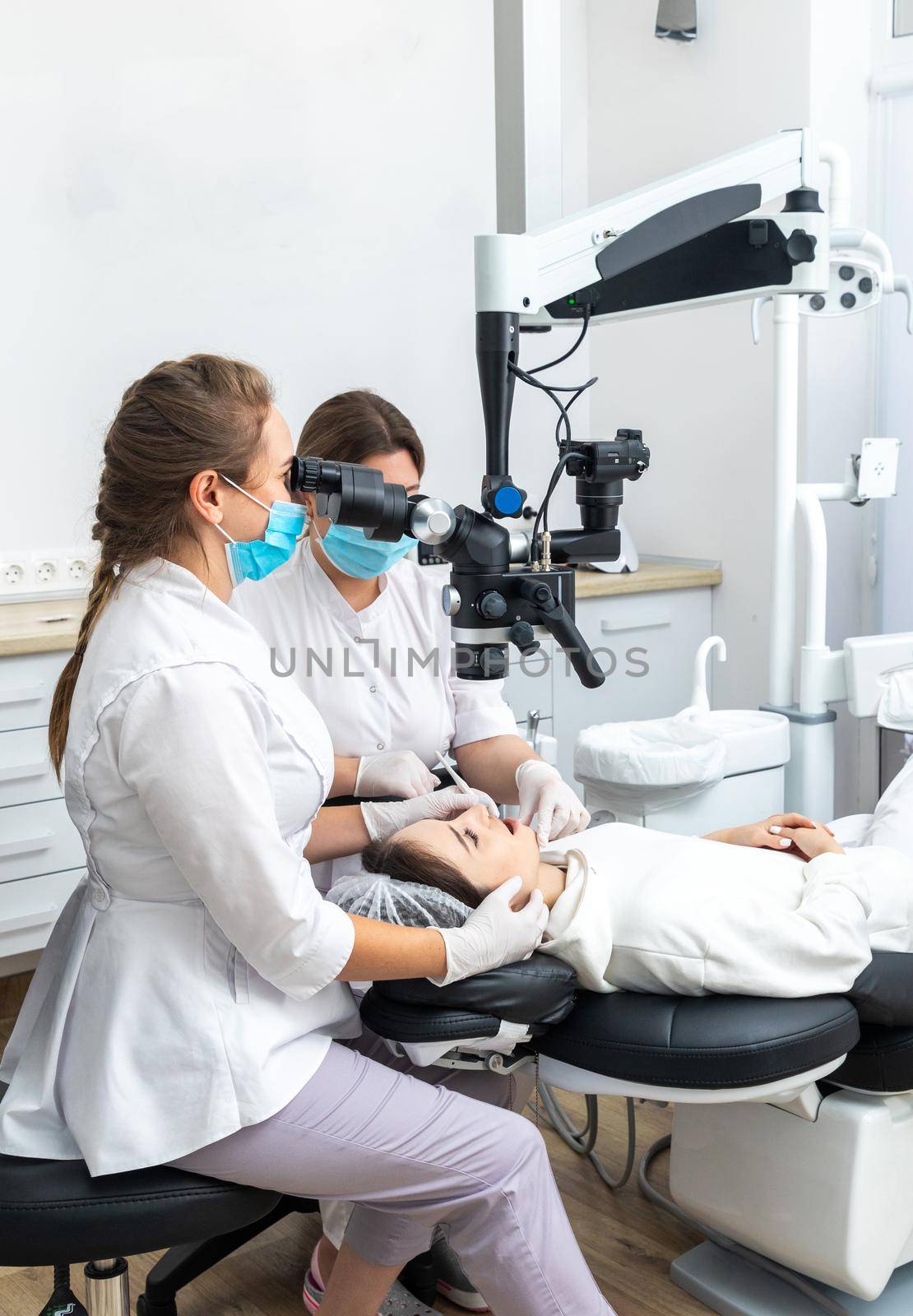 Female dentist using dental microscope treating patient teeth at dental clinic office by Mariakray