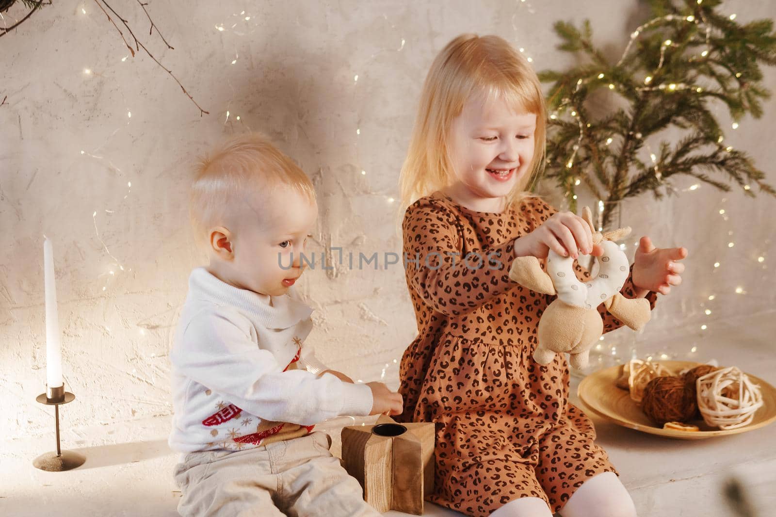Little brother and sister play on Christmas eve in a beautiful house decorated for the New Year holidays. Children are playing with a Christmas gift. Scandinavian-style interior with live fir trees and a wooden staircase.