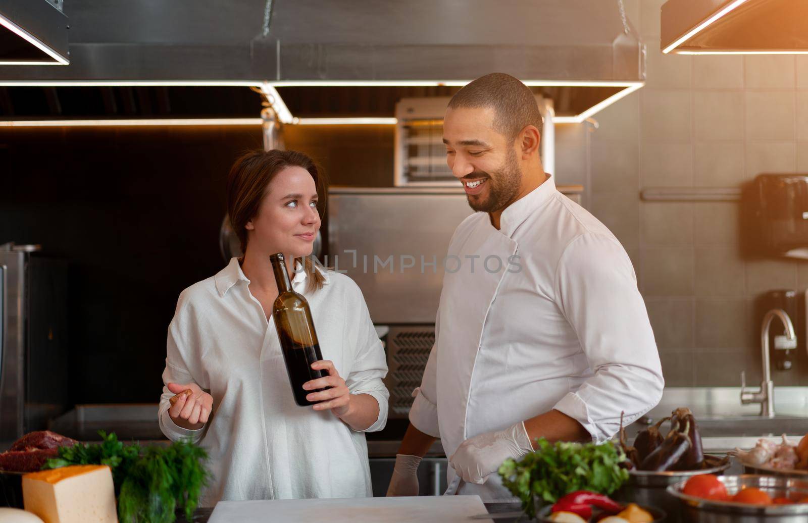 Handsome young African chef is cooking together with Caucasian girlfriend in the kitchen using red wine ingredient. A cook teaches a girl how to cook. Man and woman cooking in professional kitchen.