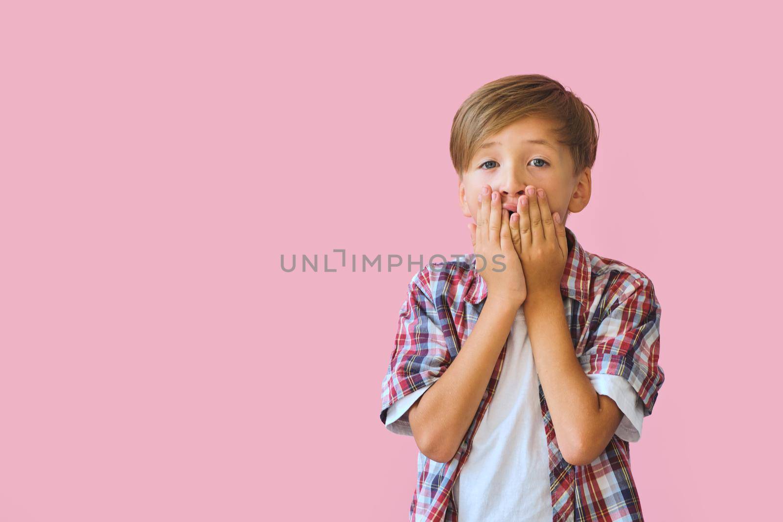 Young surprised boy dressed in jeans, a white T-shirt and a plaid shirt covers mouth with palms, isolated on pink background with copy space