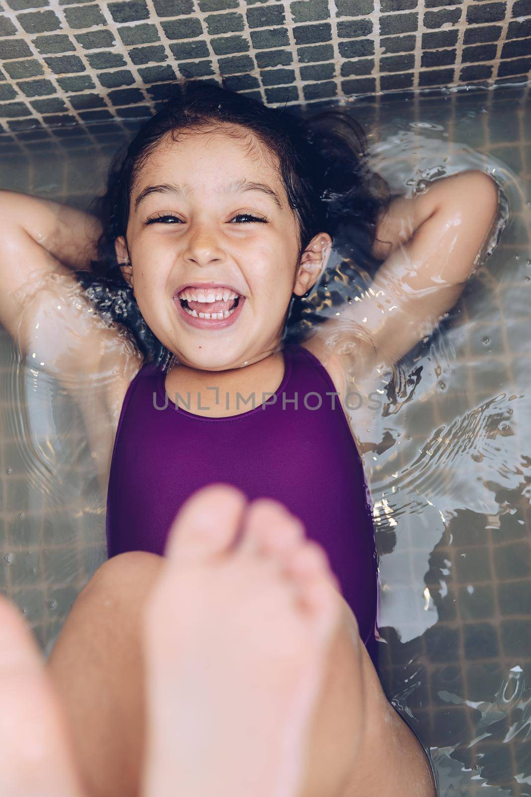 vertical photo of a playful beautiful little girl having fun and laughing lying in the tub and showing the feet while taking a bath in the bathtub, kids hygiene concept