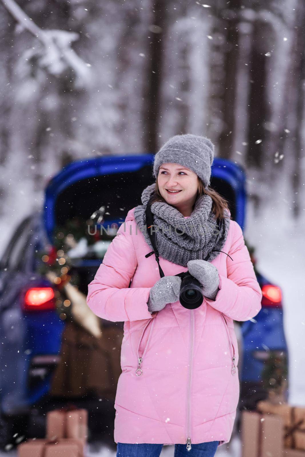 A woman in a winter snow-covered forest in the trunk of a car decorated with Christmas decor. A female photographer holds a camera in her hands.