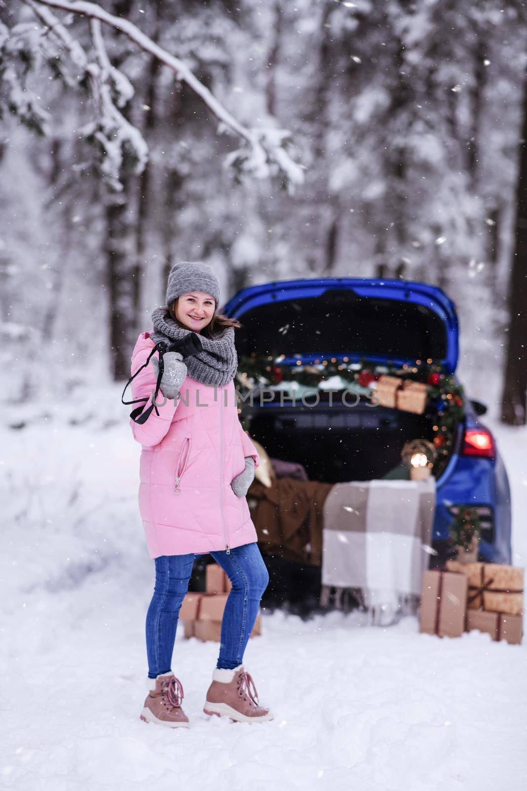 A woman in a winter snow-covered forest in the trunk of a car decorated with Christmas decor. A female photographer holds a camera in her hands.