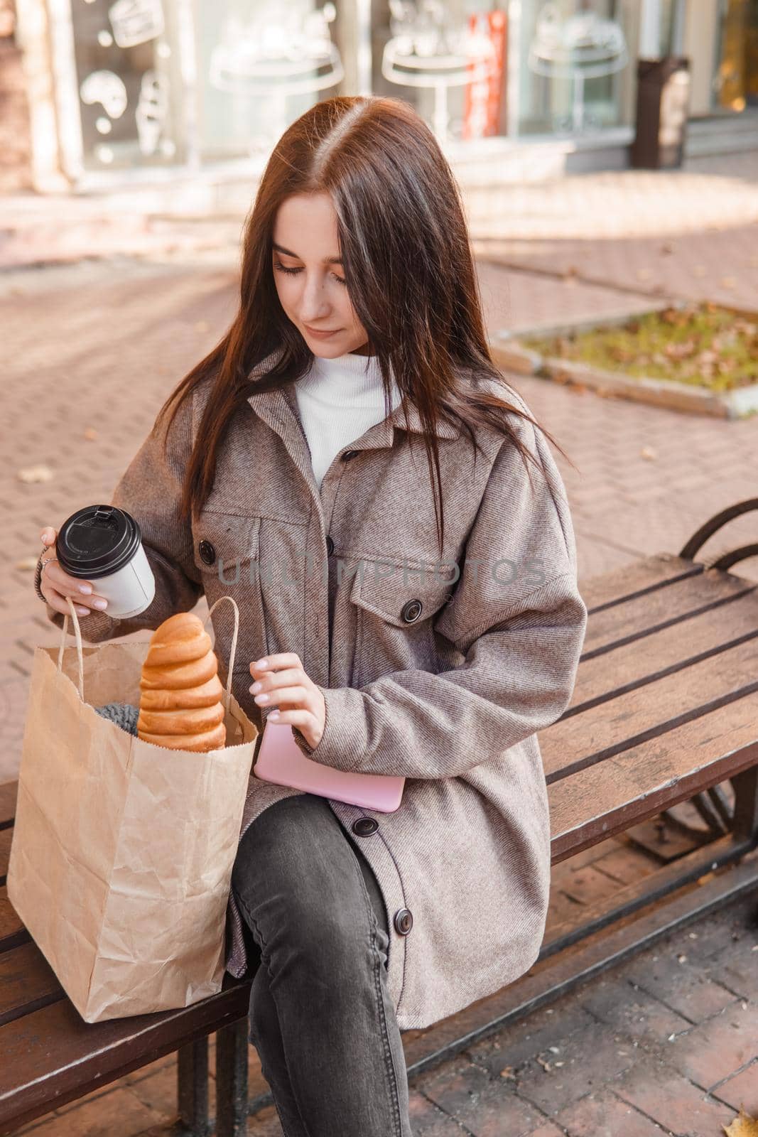A stylish brunette woman walks around the autumn city. The brunette is sitting on a bench outside with a glass of coffee and a phone, holding a craft bag with a baguette.