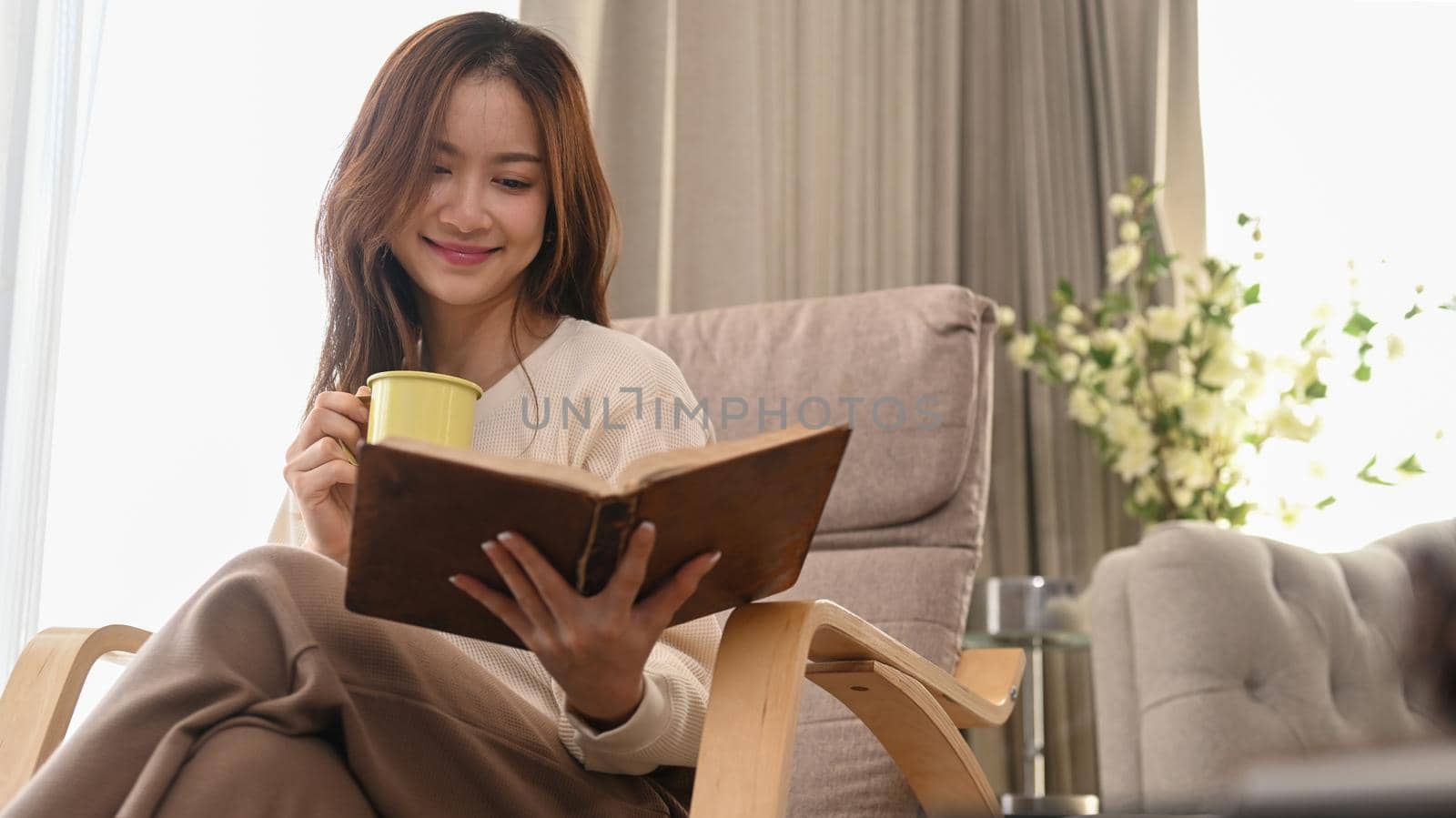 Pleasant young woman drinking herbal tea and reading book on armchair, spending leisure weekend at home.