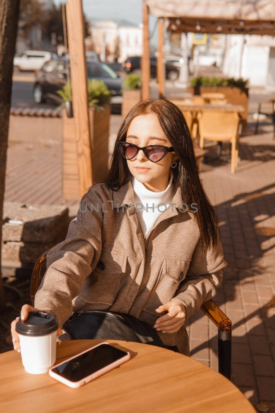 A stylish brunette woman in sunglasses is sitting at a table in an outdoor cafe with coffee and a phone. The girl is talking on the phone. Autumn walk.