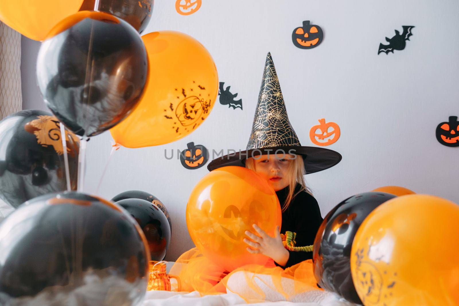 Children's Halloween - a girl in a witch hat and a carnival costume with airy orange and black balloons at home. Ready to celebrate Halloween.