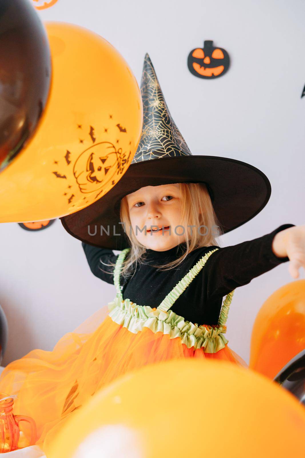 Children's Halloween - a girl in a witch hat and a carnival costume with airy orange and black balloons at home. Ready to celebrate Halloween.