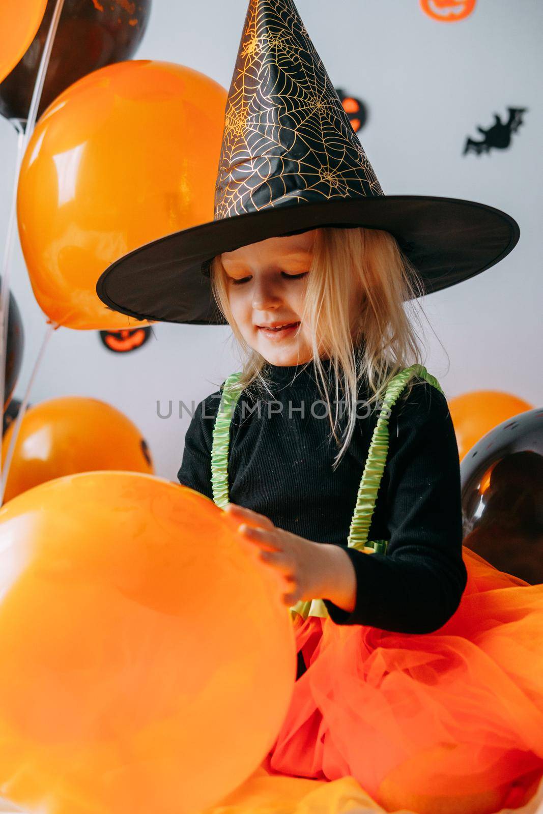 Children's Halloween - a girl in a witch hat and a carnival costume with airy orange and black balloons at home. Ready to celebrate Halloween.