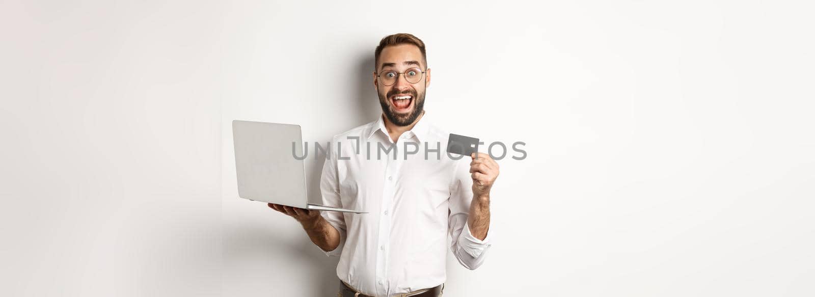 Online shopping. Handsome man showing credit card and using laptop to order in internet, standing over white background.