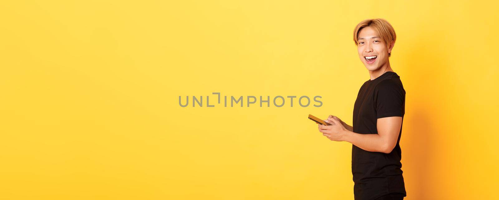 Portrait of handsome stylish asian guy in black outfit, using mobile phone and turning head at camera with satisfied smile, yellow background by Benzoix