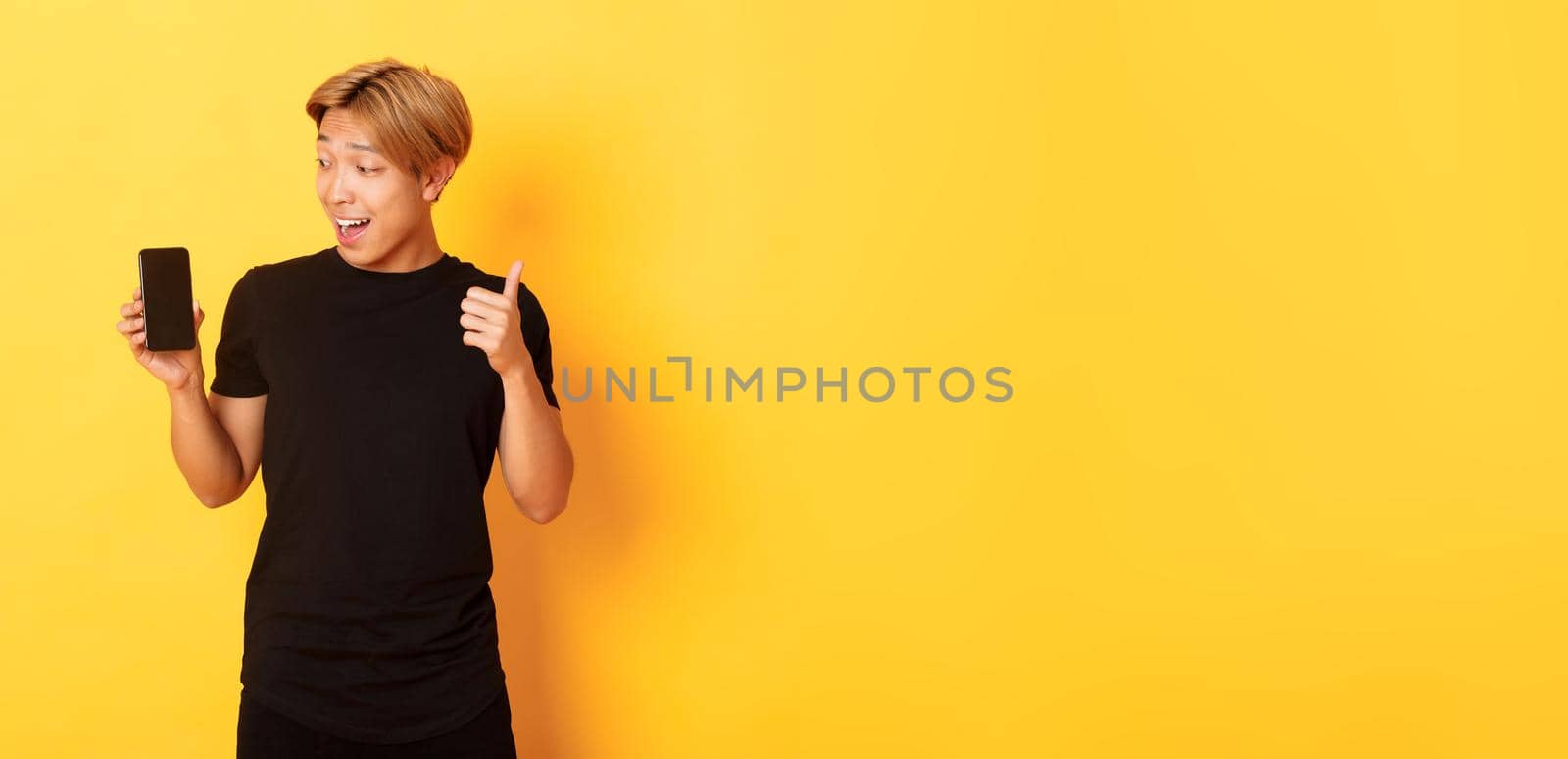 Portrait of satisfied asian guy looking at smartphone screen and showing thumbs-up in approval, standing yellow background.