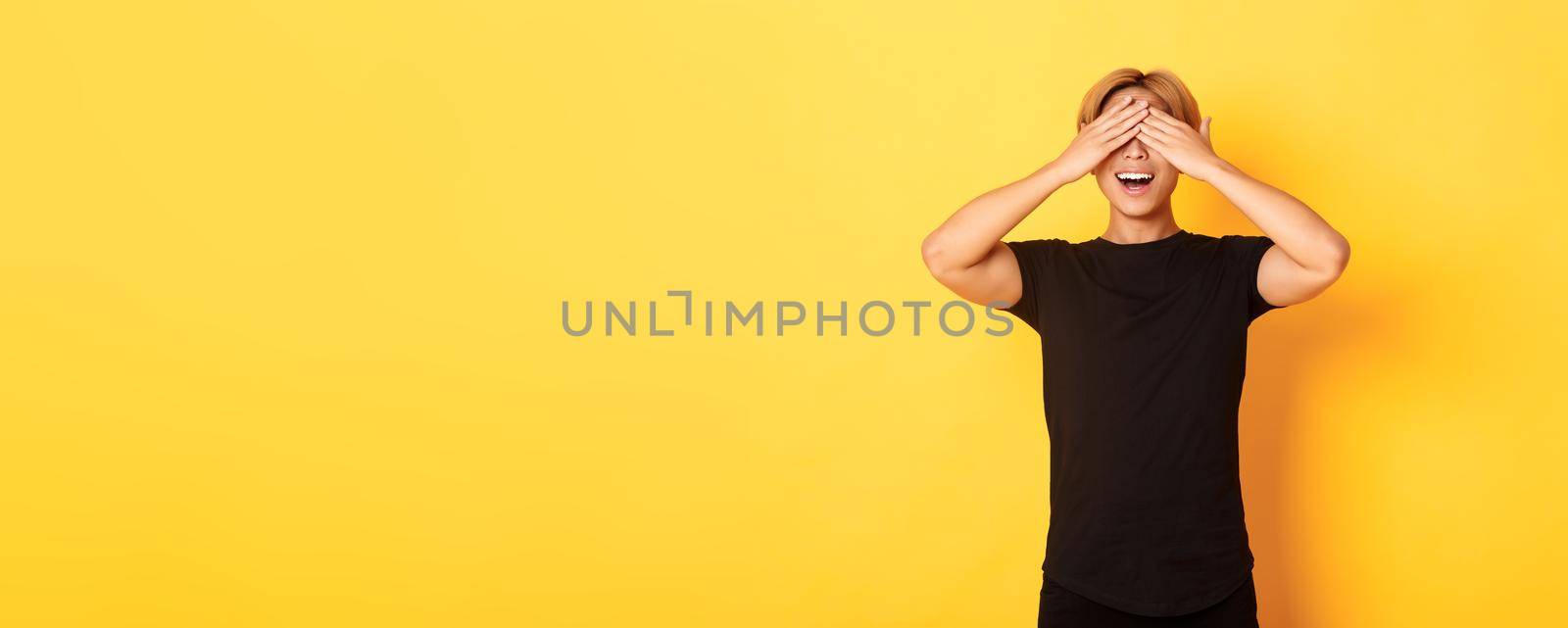 Portrait of amused and excited smiling blond guy shut eyes with palms, waiting for surprise, standing yellow background.