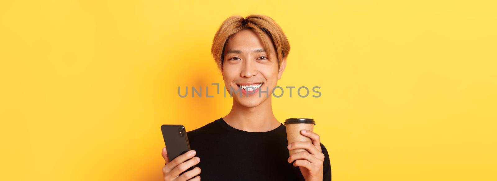 Close-up of handsome young asian guy smiling happy, using smartphone and drinking takeaway coffee, standing over yellow background by Benzoix