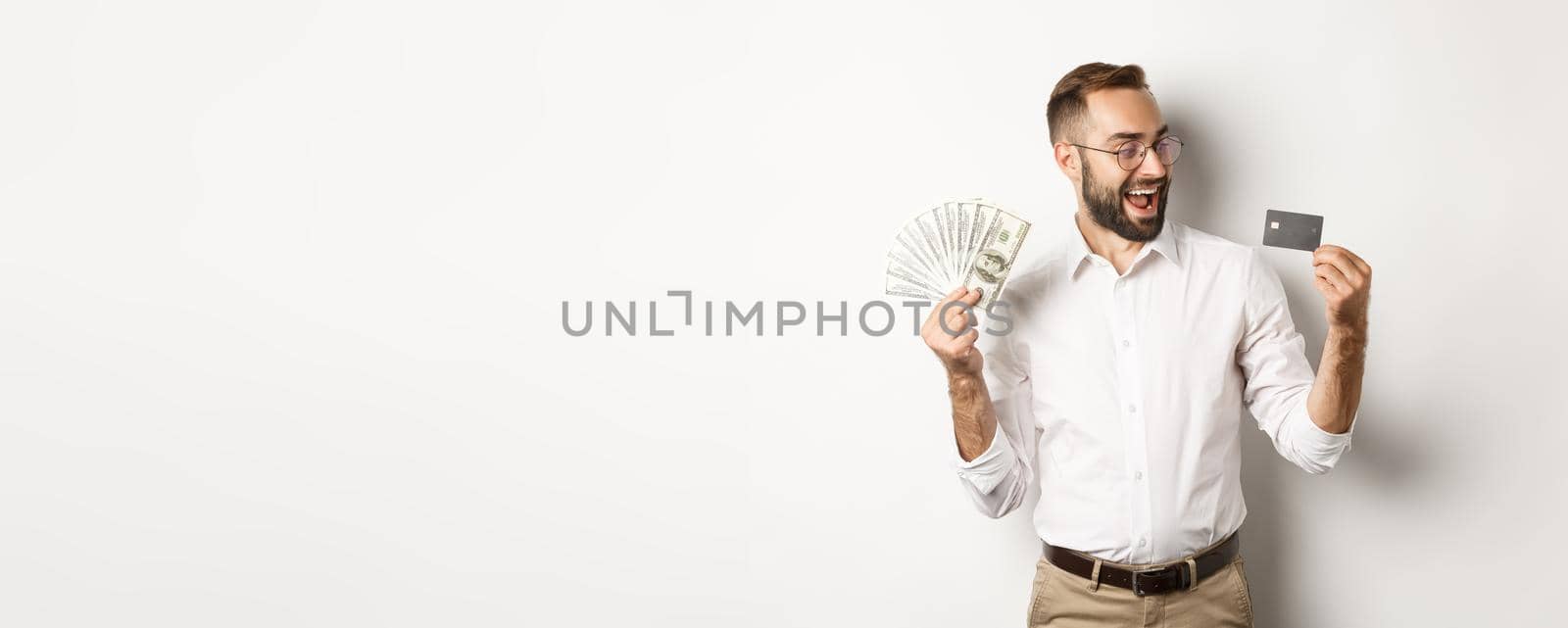 Excited businessman holding money and looking at credit card, standing over white background by Benzoix