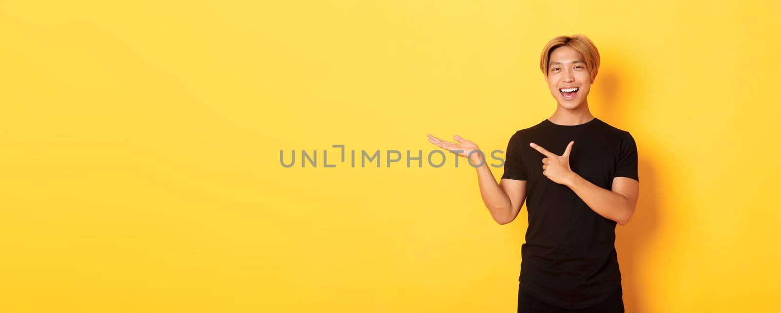 Portrait of handsome smiling asian guy holding something on hand and pointing finger at it, standing yellow background pleased.