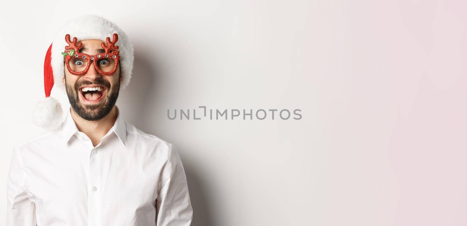 Close-up of excited bearded man in christmas glasses and santa hat looking amazed at promo offer, winter holidays advertisement concept, white background.