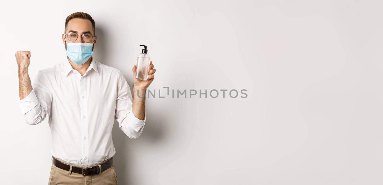 Covid-19, social distancing and quarantine concept. Cheerful manager in medical mask showing hand sanitizer, standing over white background.