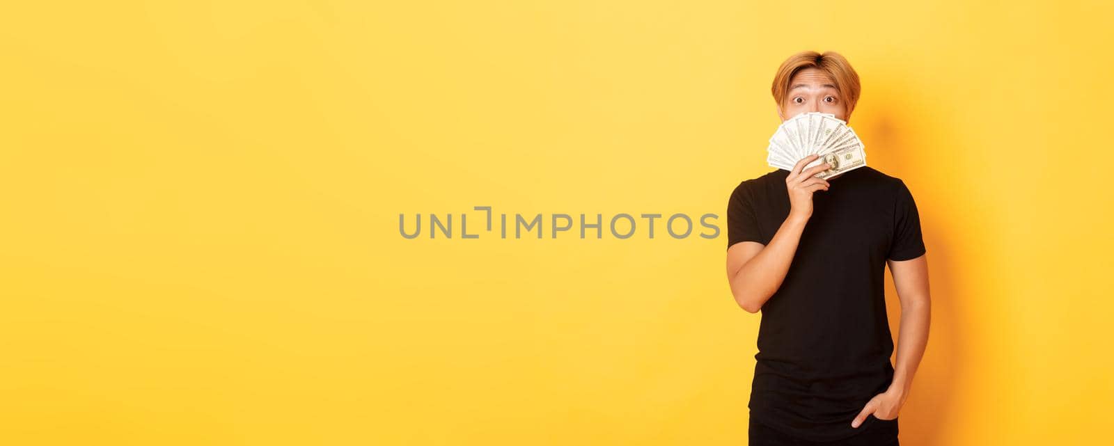 Portrait of lucky happy asian guy looking surprised and showing money, standing yellow background.