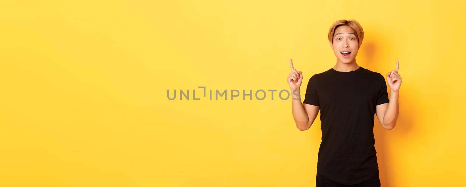 Portrait of surprised and excited asian guy looking amused, pointing fingers up, yellow background.