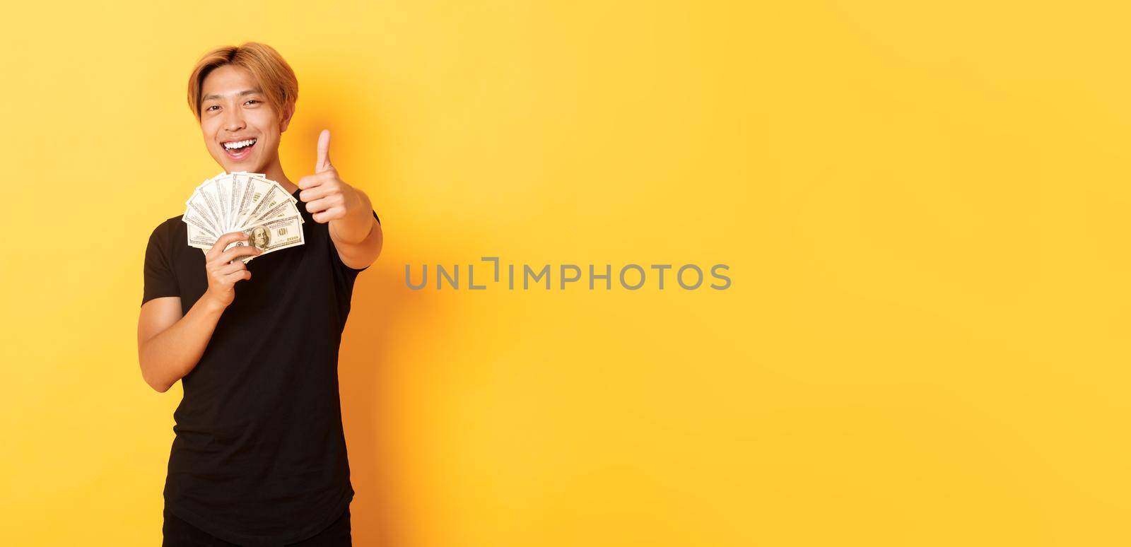 Portrait of handsome confident smiling asian guy showing thumbs-up and holding money, guarantee something, standing yellow background by Benzoix