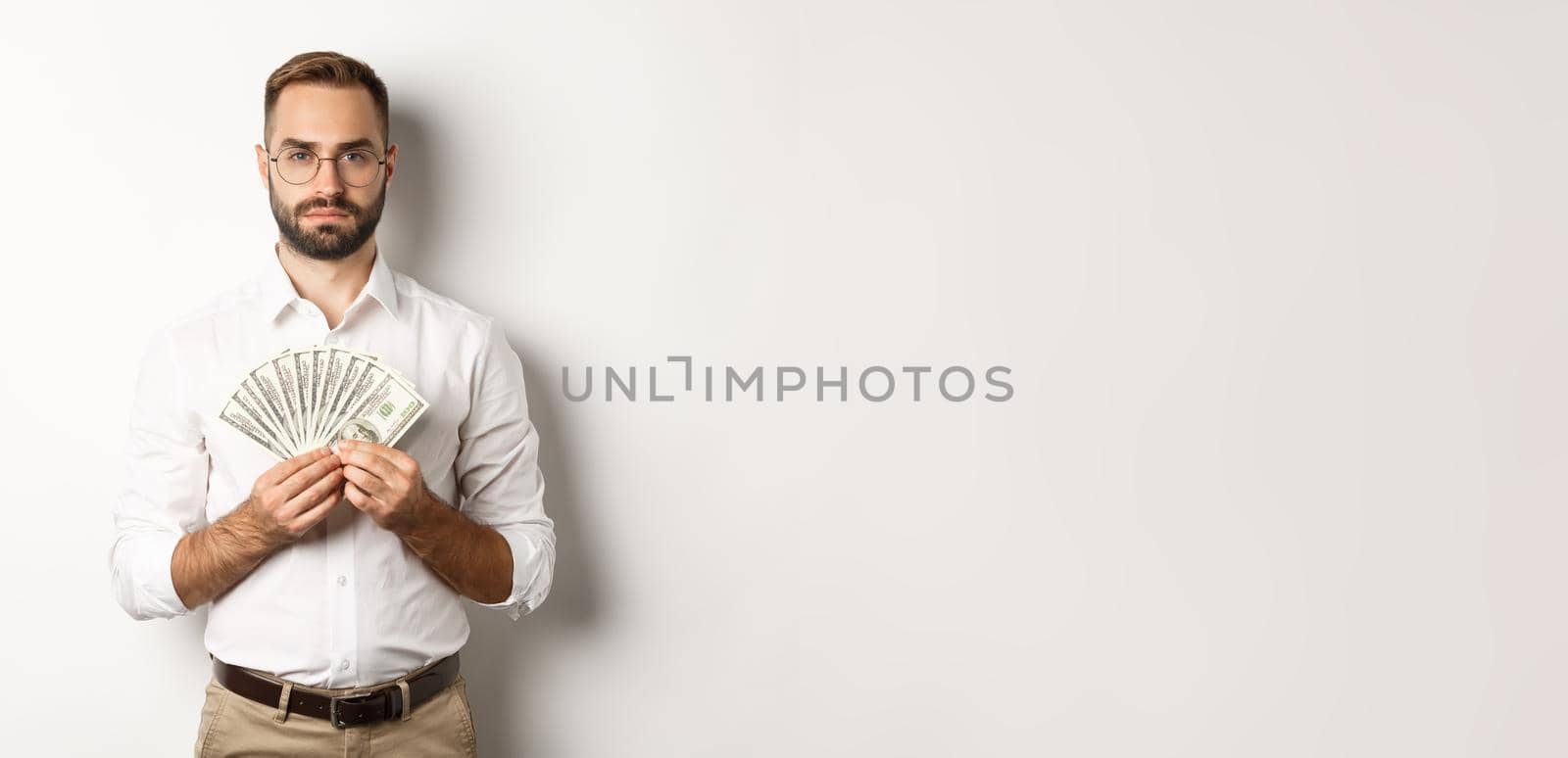 Serious businessman holding money, showing dollars, standing over white background by Benzoix