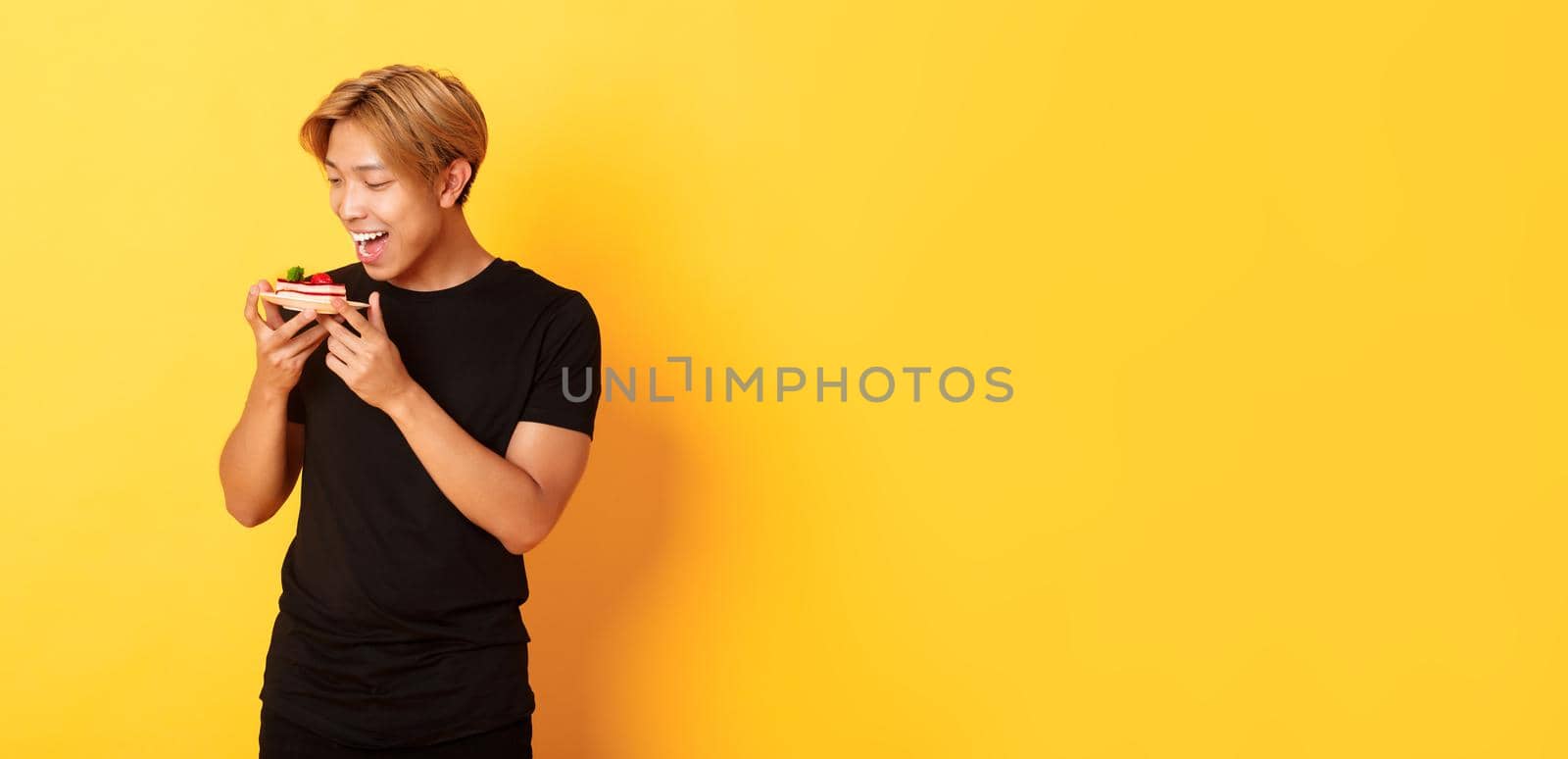 Joyful handsome asian guy smiling and looking tempted at cake while making bite, standing yellow background.