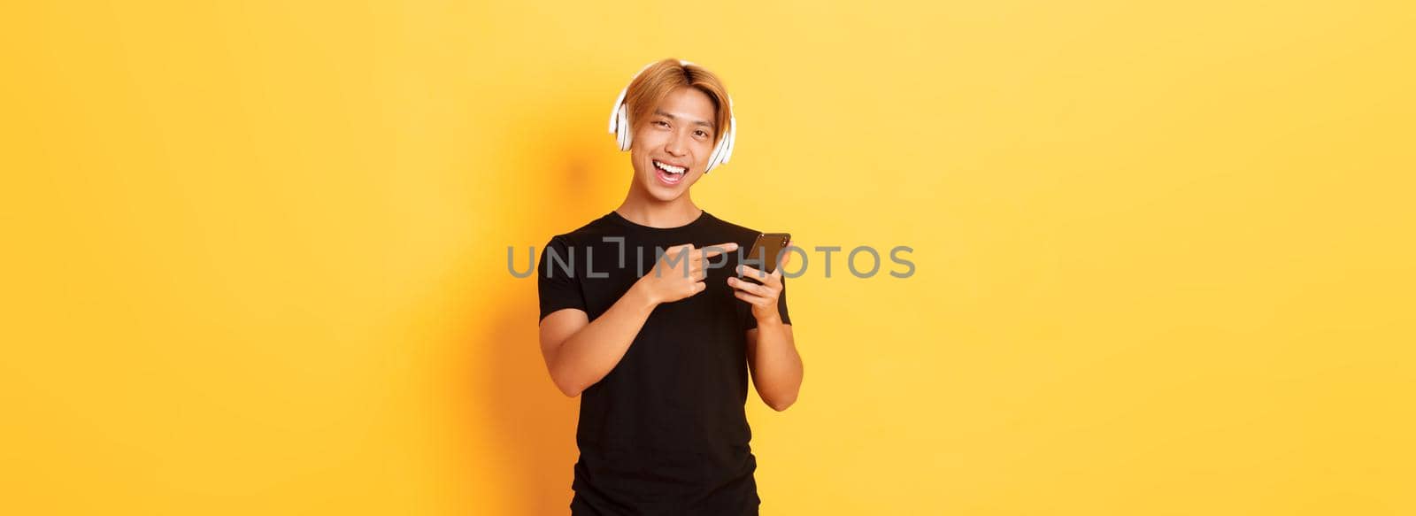 Joyful satisfied asian handsome guy, listening music or good podcast in headphones, pointing finger at smartphone with pleased smile, yellow background by Benzoix