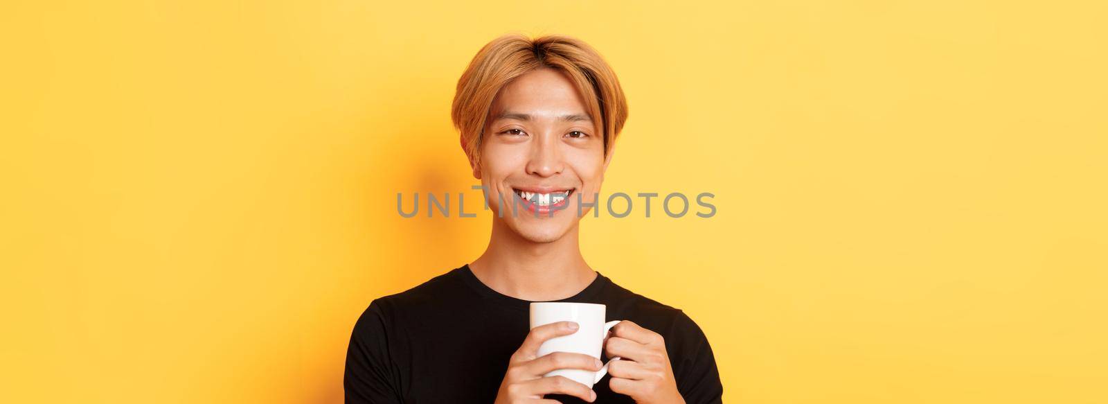 Close-up of happy satisfied asian smiling guy, holding mug with coffee, drinking and looking pleased, standing over yellow background.