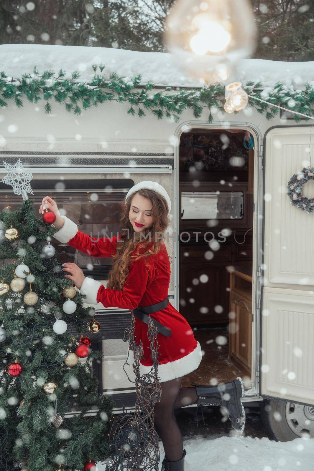 Young woman in santa costume decorates the Christmas tree at winter campsite getting ready for the new year. New year celebration concept