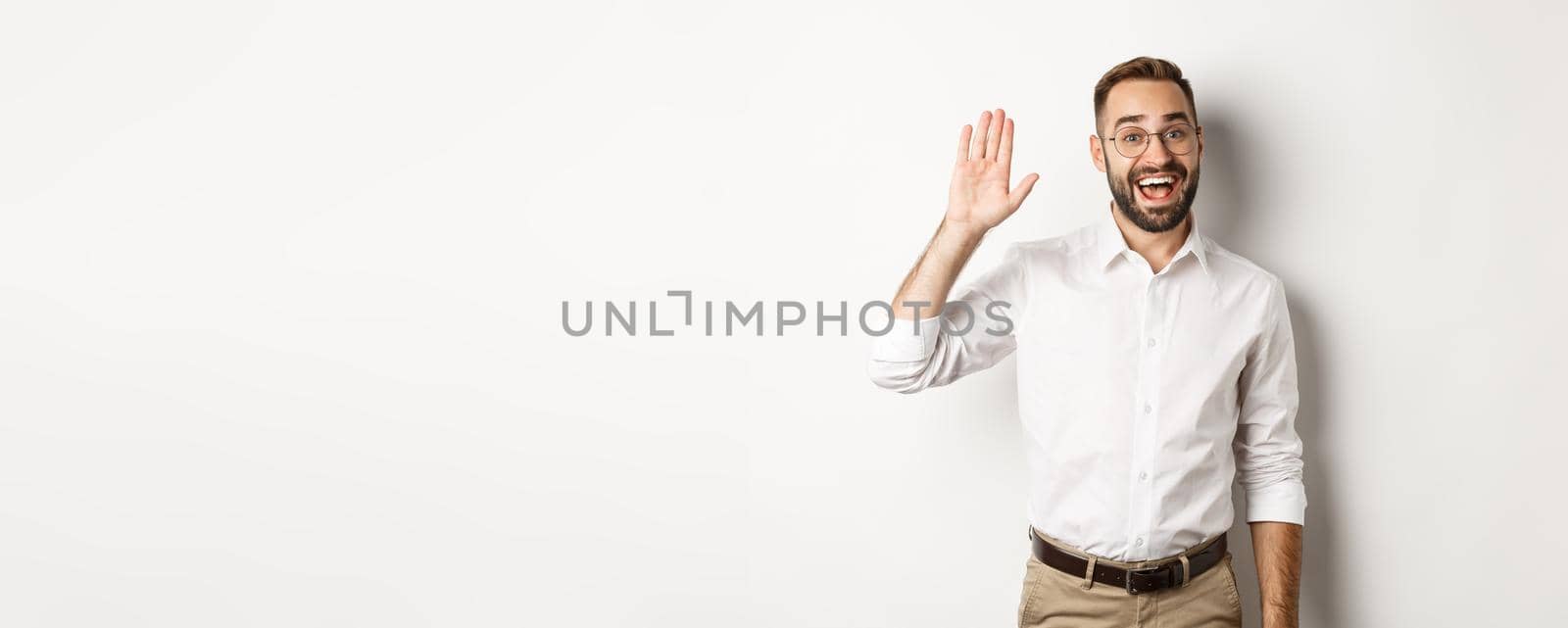 Friendly smiling man in glasses saying hello, waving hand in greeting, standing over white background.