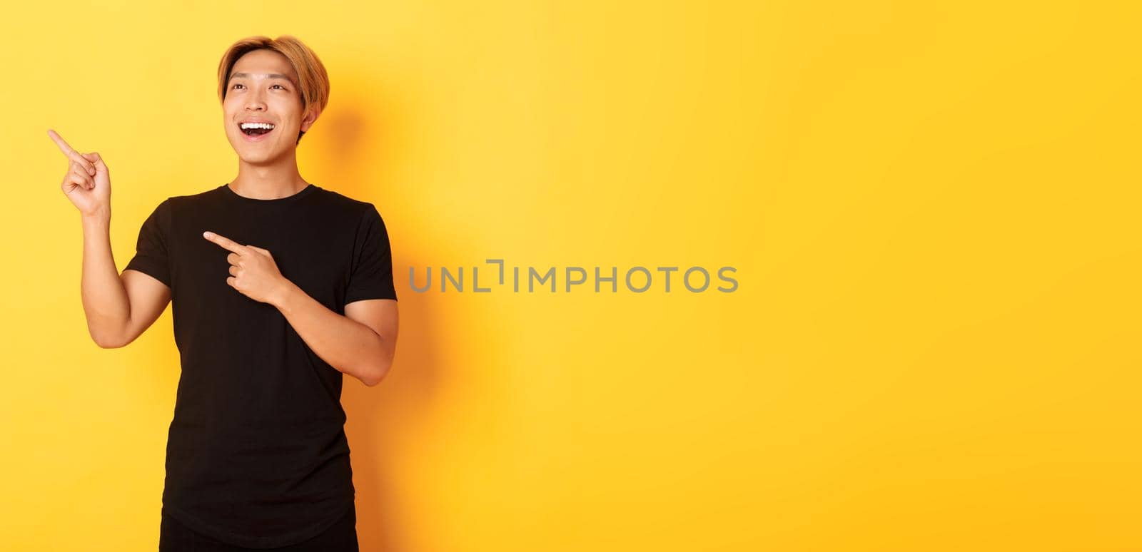 Portrait of pleased handsome asian guy in black t-shirt, pointing fingers and looking upper left corner with satisfied smile, yellow background.