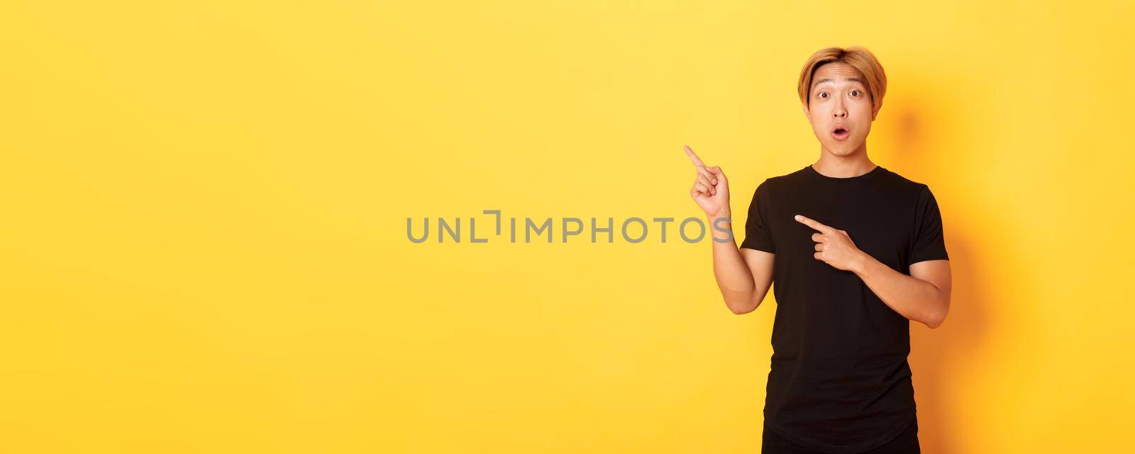 Portrait of impressed and excited handsome asian guy in black t-shirt, react to your logo, pointing fingers upper left corner.