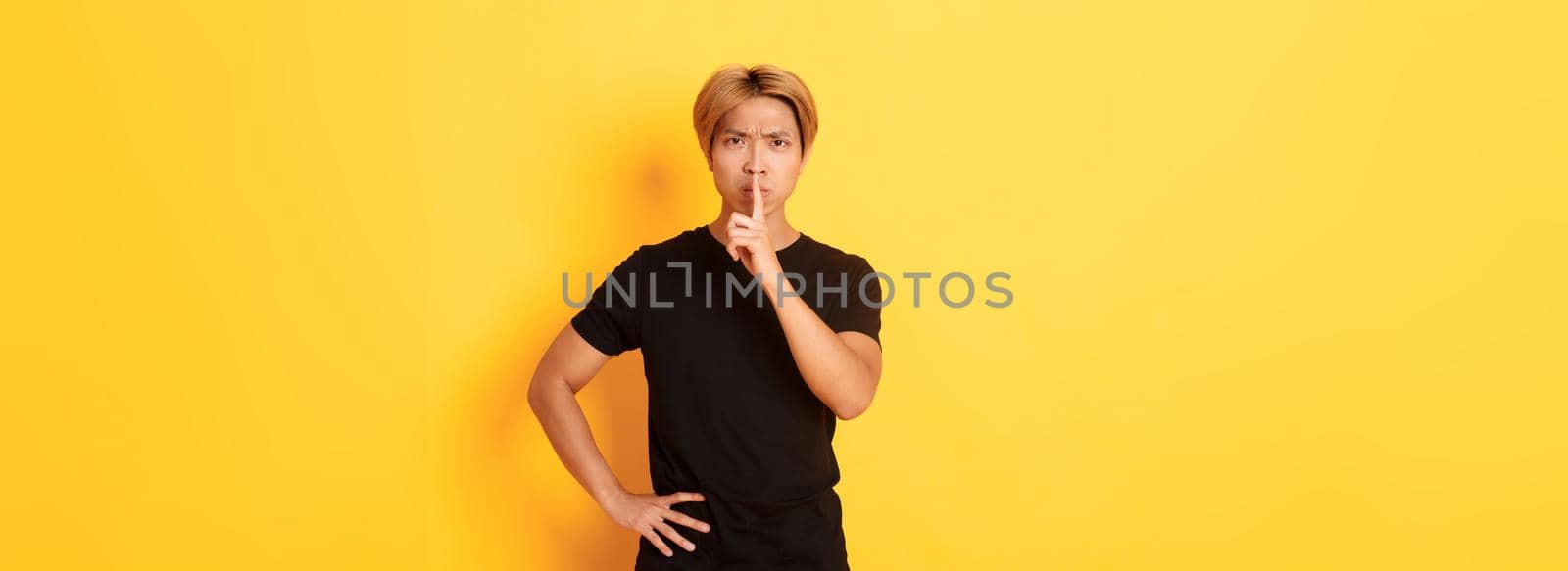 Portrait of angry asian man frowning, shushing at camera disappointed, standing yellow background by Benzoix