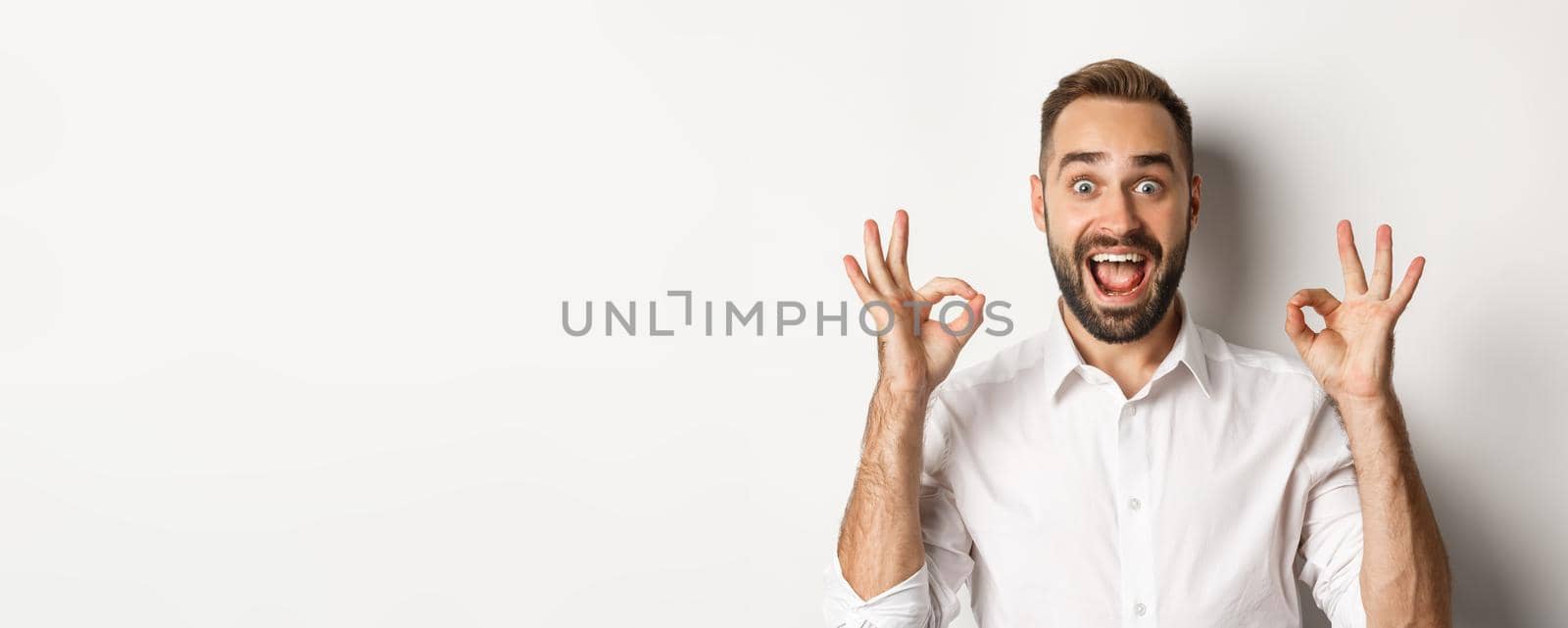 Close-up of amazed caucasian guy agreeing, showing okay signs and looking impressed, recommending product, white background.